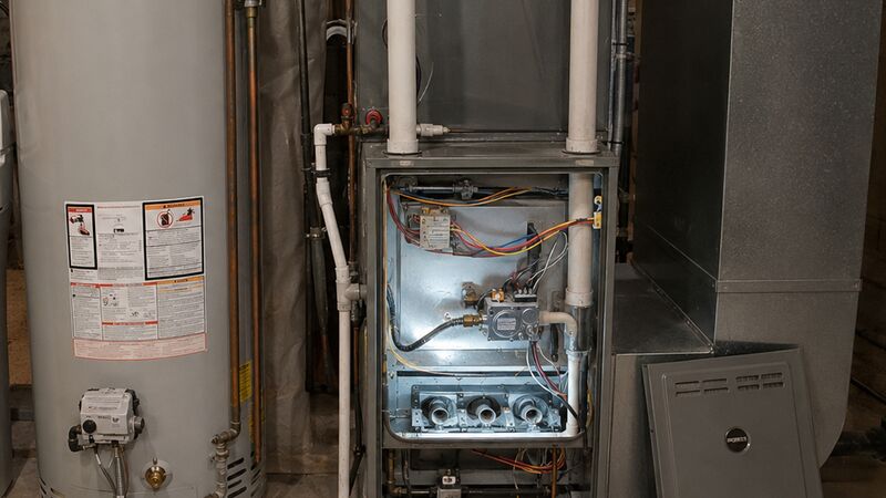 Residential gas furnace with access panel removed exposing burners and controls, alongside a water heater and a technician's open tool bag